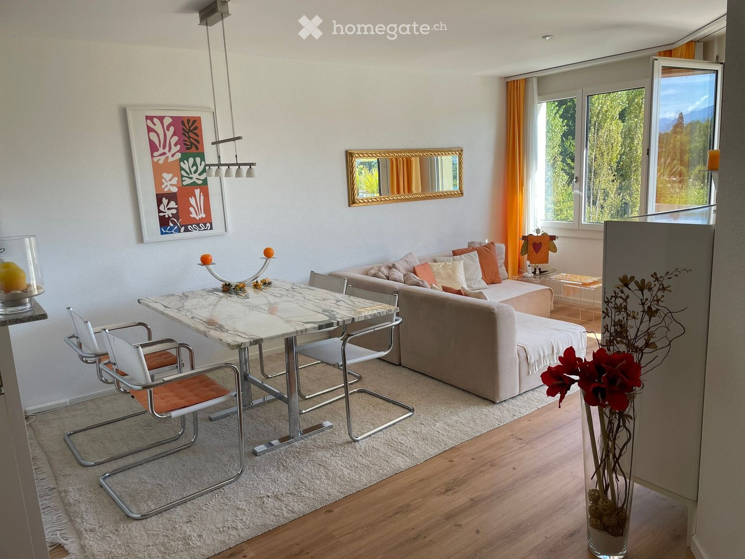 Modern living room with a beige sofa, white and orange cushions, a marble dining table with four chairs, a chandelier, a vase with red flowers, and a mirror on the wall.