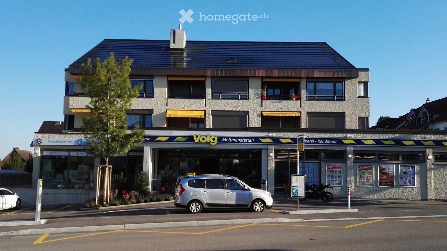 Two-story commercial building with storefronts labeled "Volg Mettmenstellen" and "Bäckerei - Konditorei." Features large windows, yellow awnings, and a small tree in front.