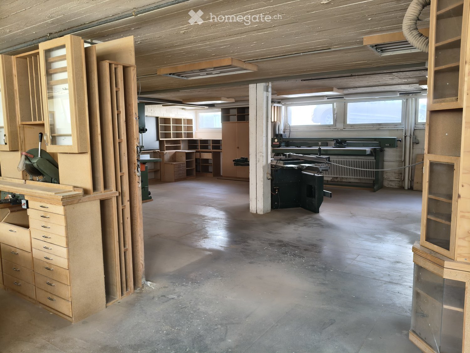 Empty workshop with exposed wooden beams, concrete floor, and various wooden furniture pieces and tools stacked around the space.