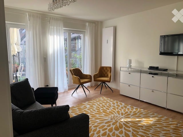Modern living room with a grey sofa, two yellow armchairs, a coffee table, a television mounted on the wall, and a white cabinet. The room features large windows with white curtains and a patterned rug on the wooden floor.