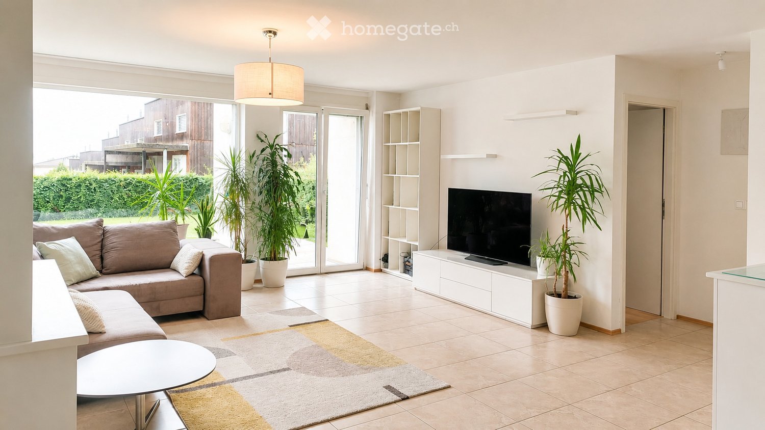 Bright living room with large sliding glass doors leading to a patio, a beige sectional sofa, a white coffee table, a flat-screen TV on a white entertainment unit, and several potted plants.
