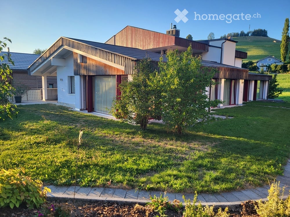 Modern two-story house with a combination of white and wooden exterior, large windows, and a well-maintained lawn with shrubs and trees.