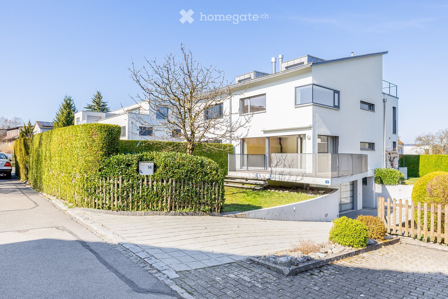 Modern white house with glass windows and a balcony, surrounded by hedges and a driveway.