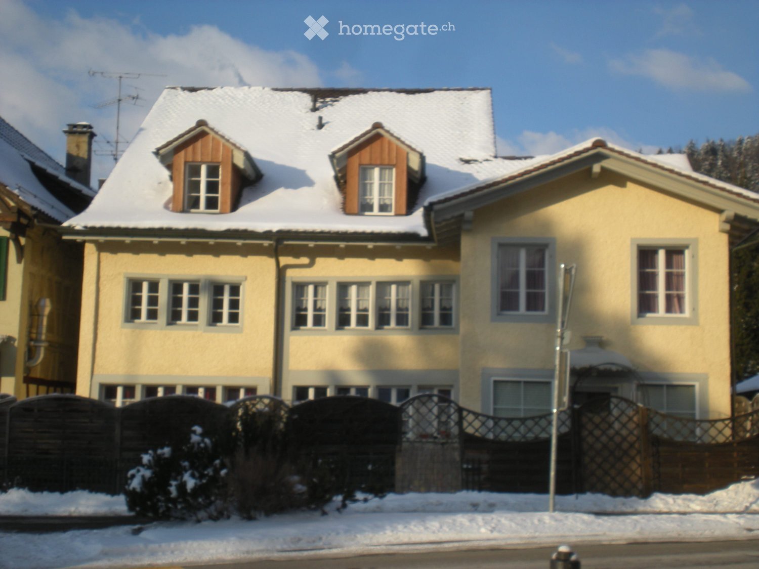 Two-story yellow house with snow-covered roof and dormer windows, surrounded by a wooden fence and snow-covered bushes.