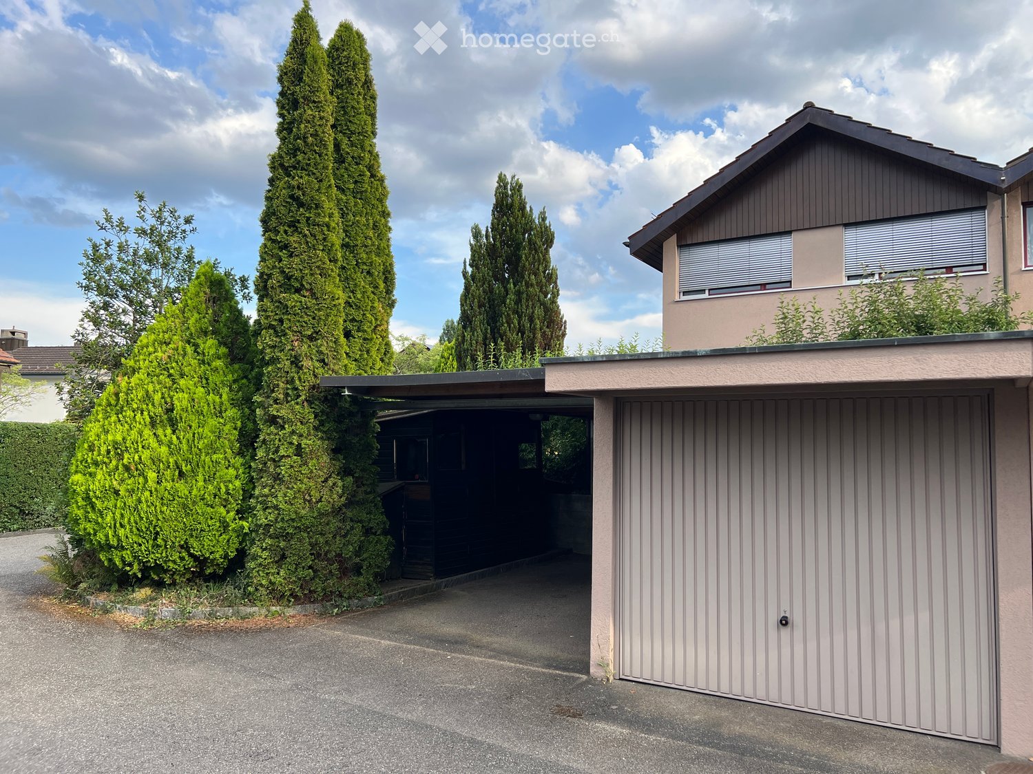 Two-story house with a garage, surrounded by tall evergreen trees and a clear driveway.