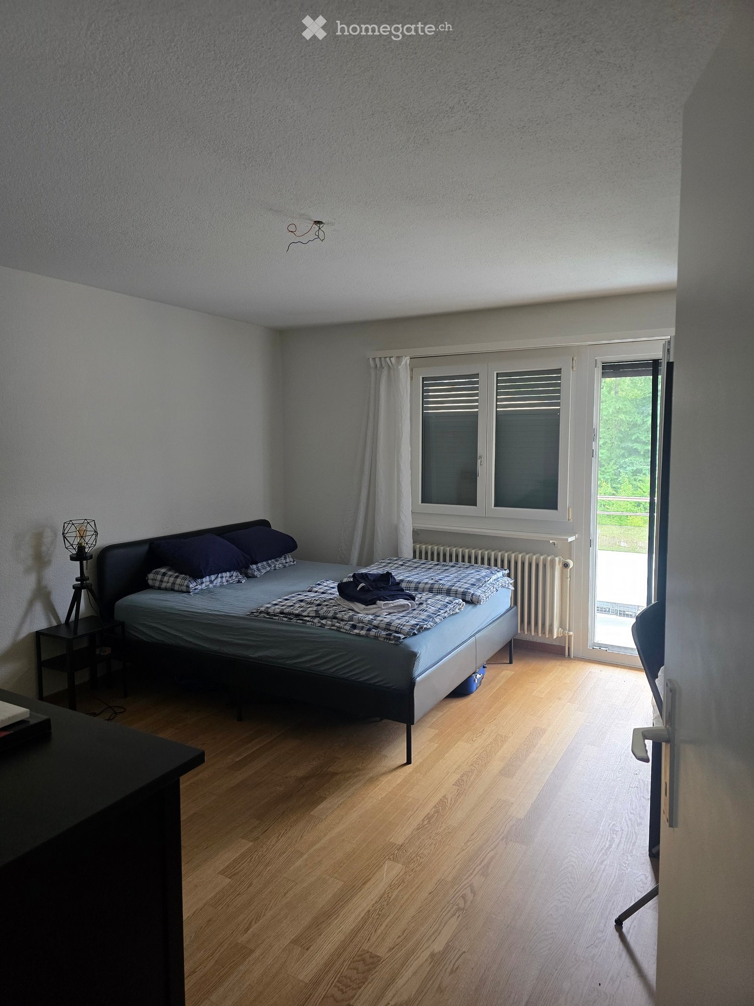 Bedroom with wooden flooring, a double bed with pillows and a blanket, a nightstand with a lamp, and a chair near the window.