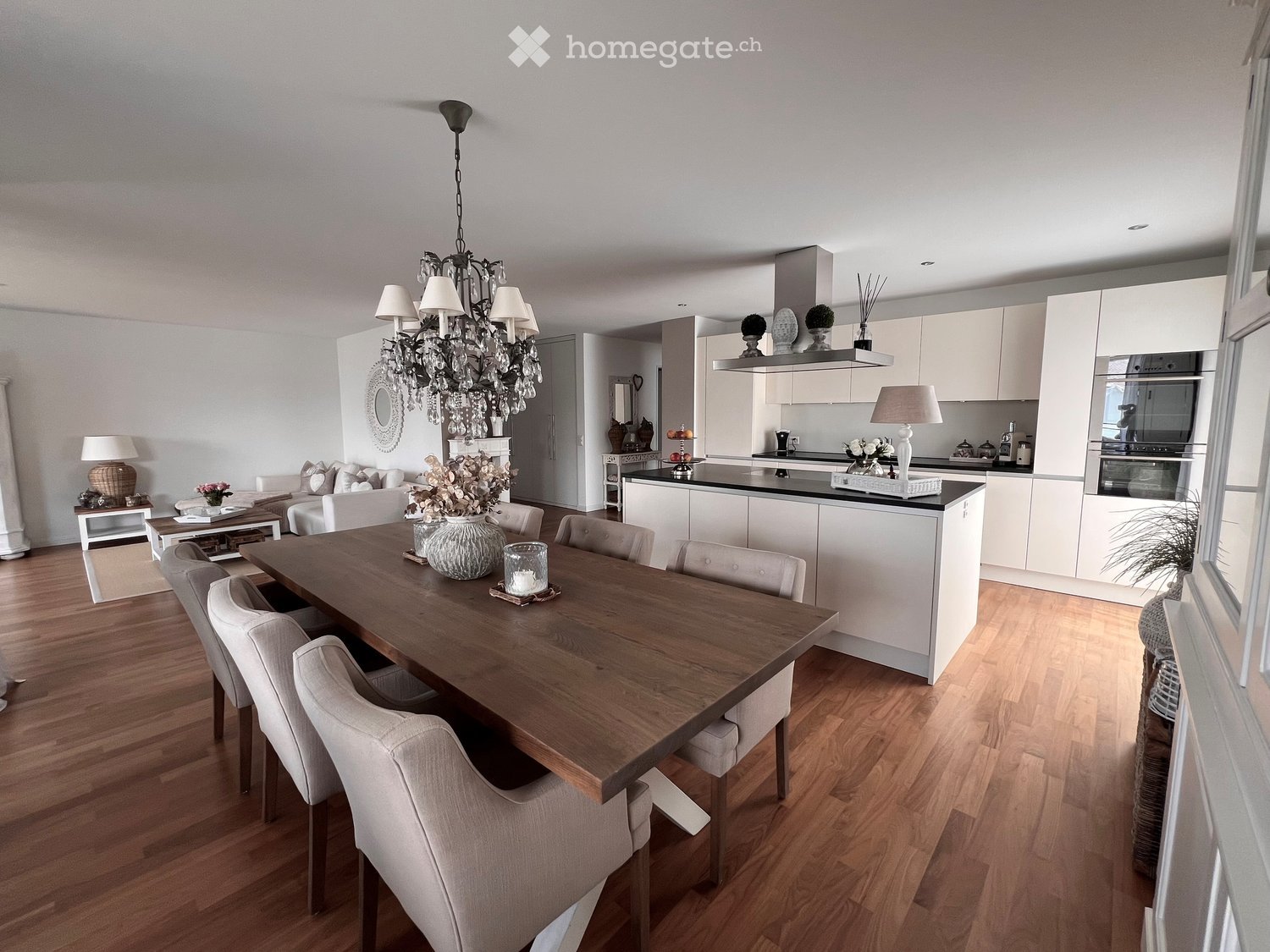 Open-plan living and dining area with a wooden dining table, six chairs, a sofa, coffee table, chandelier, and a modern kitchen with white cabinets and stainless steel appliances.