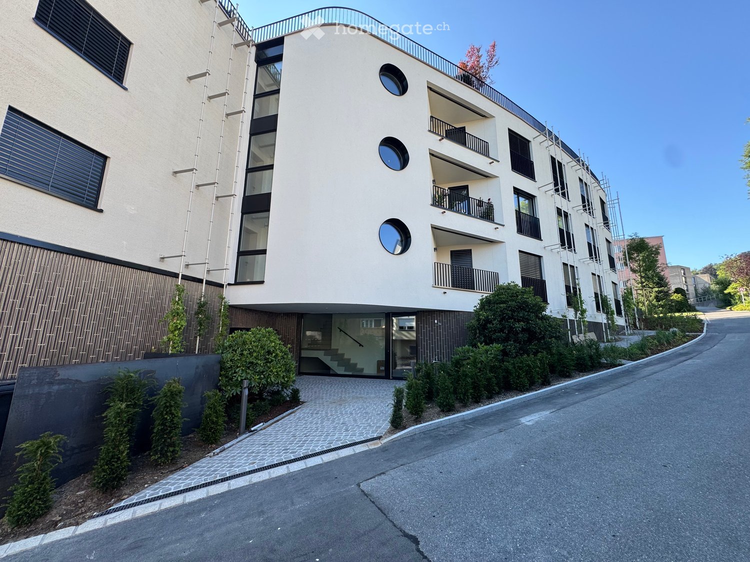 Modern multi-story residential building with balconies, large windows, and a curved roofline. Features include a raised ground floor entrance, greenery, and a paved driveway.