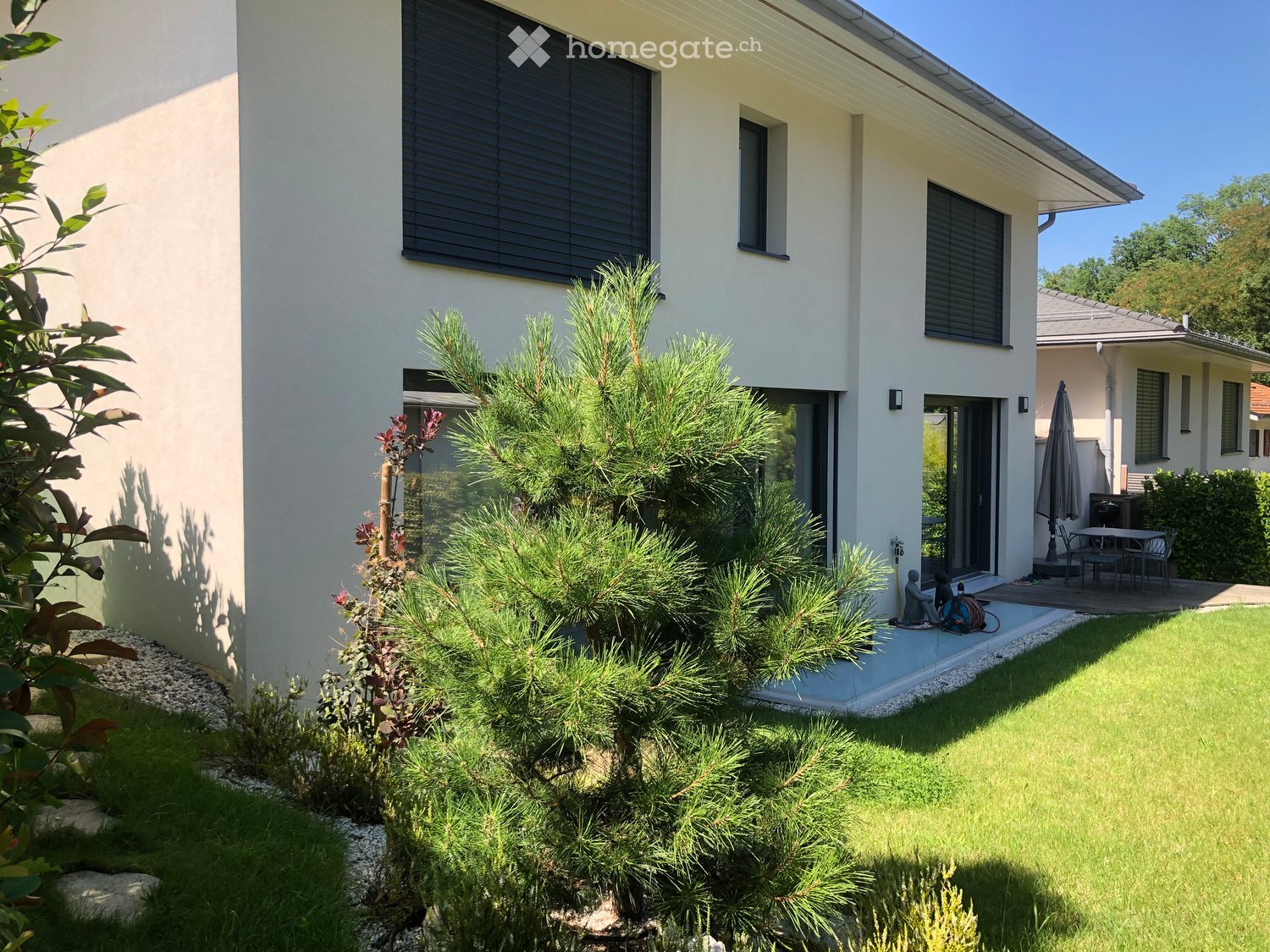 Well-maintained garden with green lawn, shrubs, and a small patio area with outdoor furniture and an umbrella.