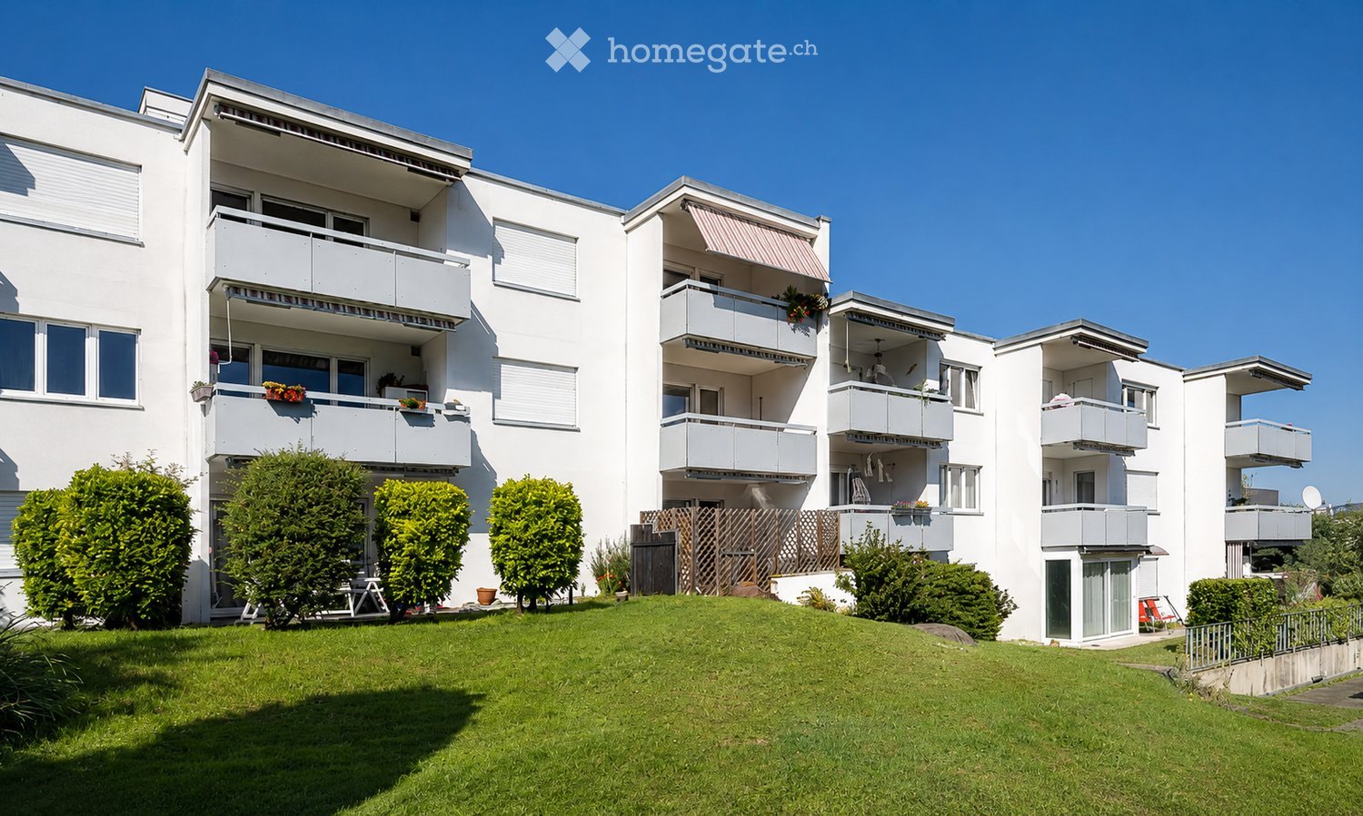 Modern white apartment building with multiple balconies, greenery, and a well-maintained lawn in front.