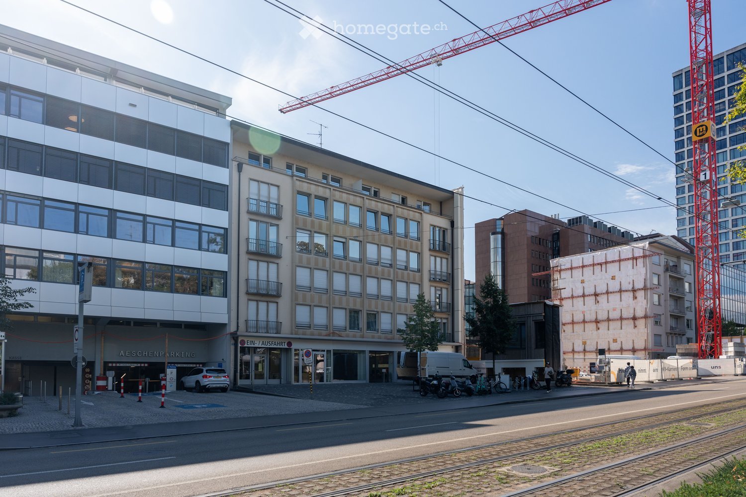 A multi-story building with a modern facade, featuring large windows and balconies. The ground floor includes a parking area with a sign reading "AESCHENPARKING" and several parked vehicles, including a white van and a white car. The surrounding area incl