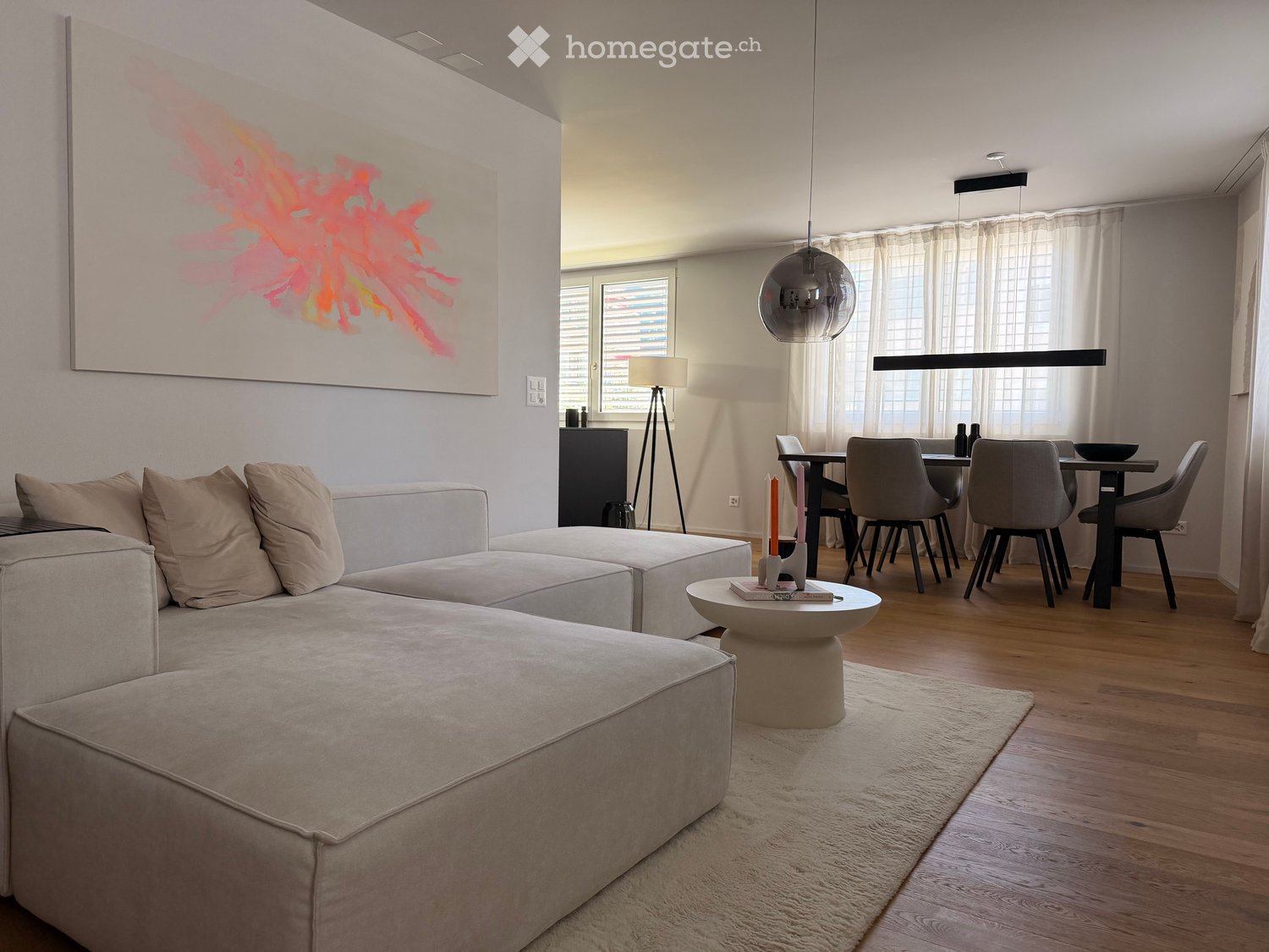 Modern living and dining area with a sectional sofa, coffee table, dining table with chairs, pendant lights, and a large abstract painting on the wall.