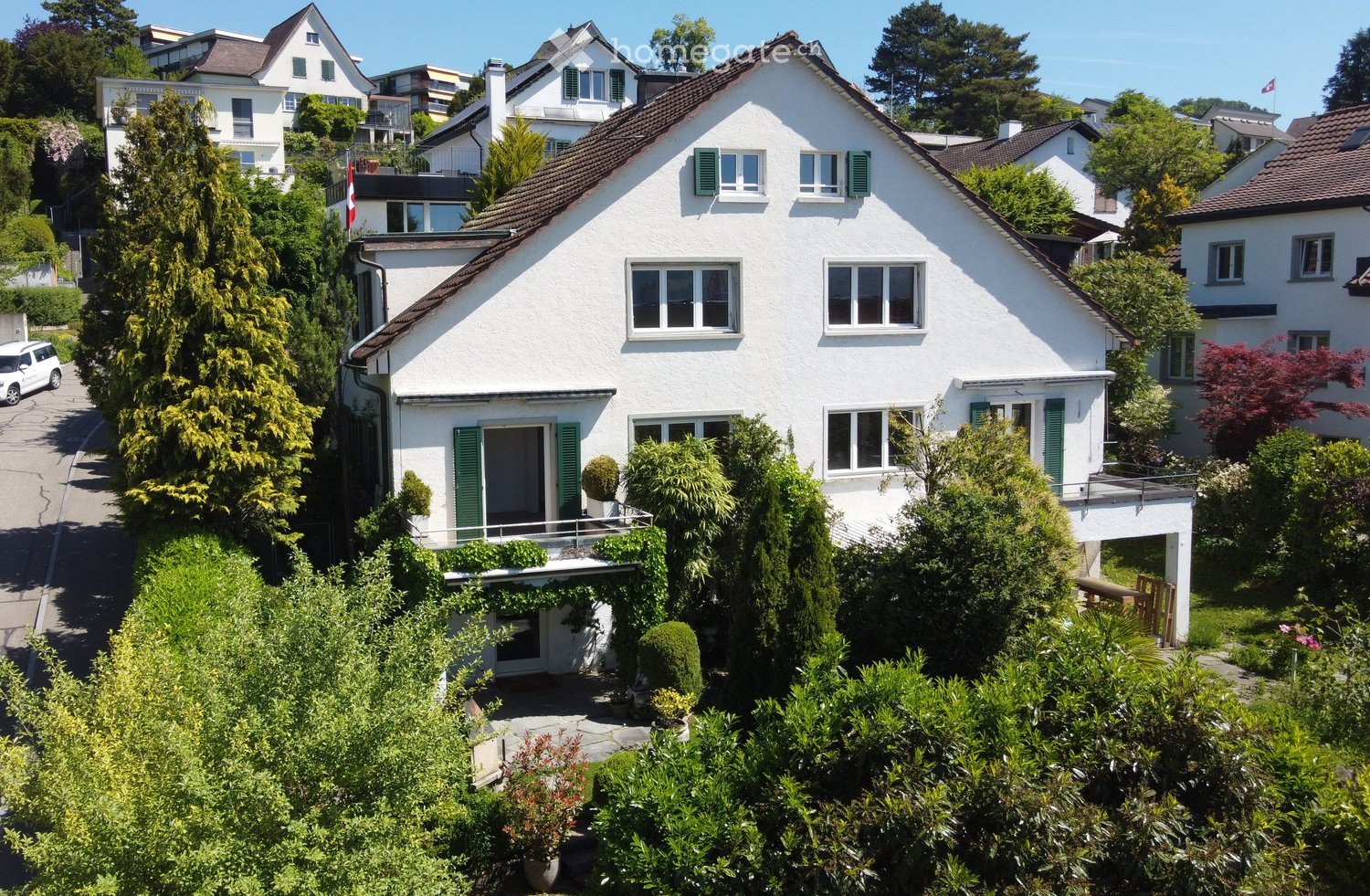 A two-story white house with green shutters, surrounded by lush greenery and trees, with a driveway and neighboring houses visible in the background.
