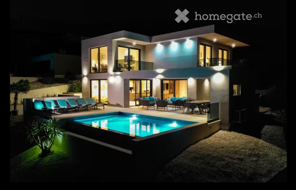 Modern two-story house with large windows, illuminated exterior, swimming pool, outdoor seating area, and lounge chairs.