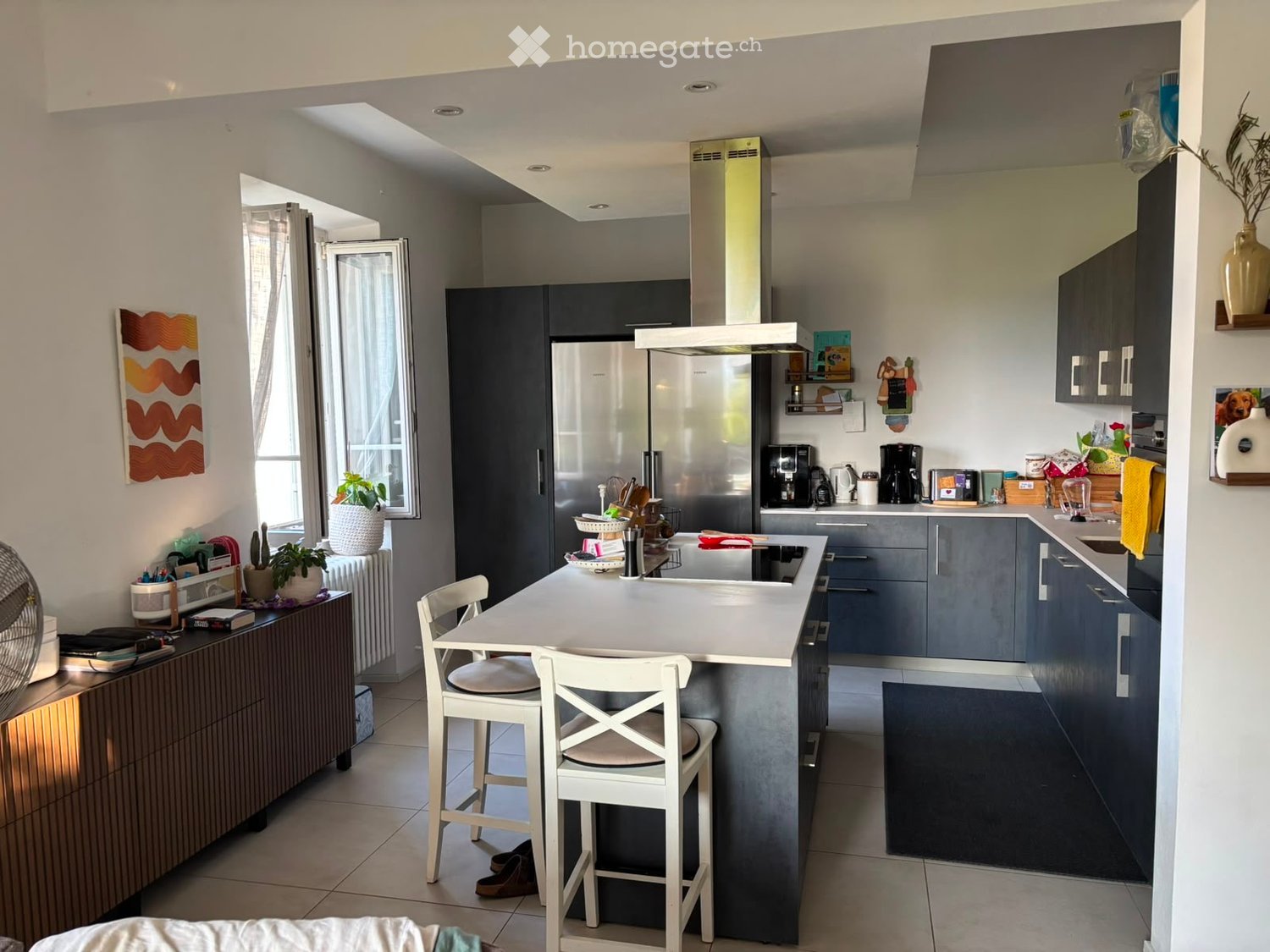 Modern kitchen with grey cabinets, stainless steel appliances, island with bar stools, and various kitchen items on counters.