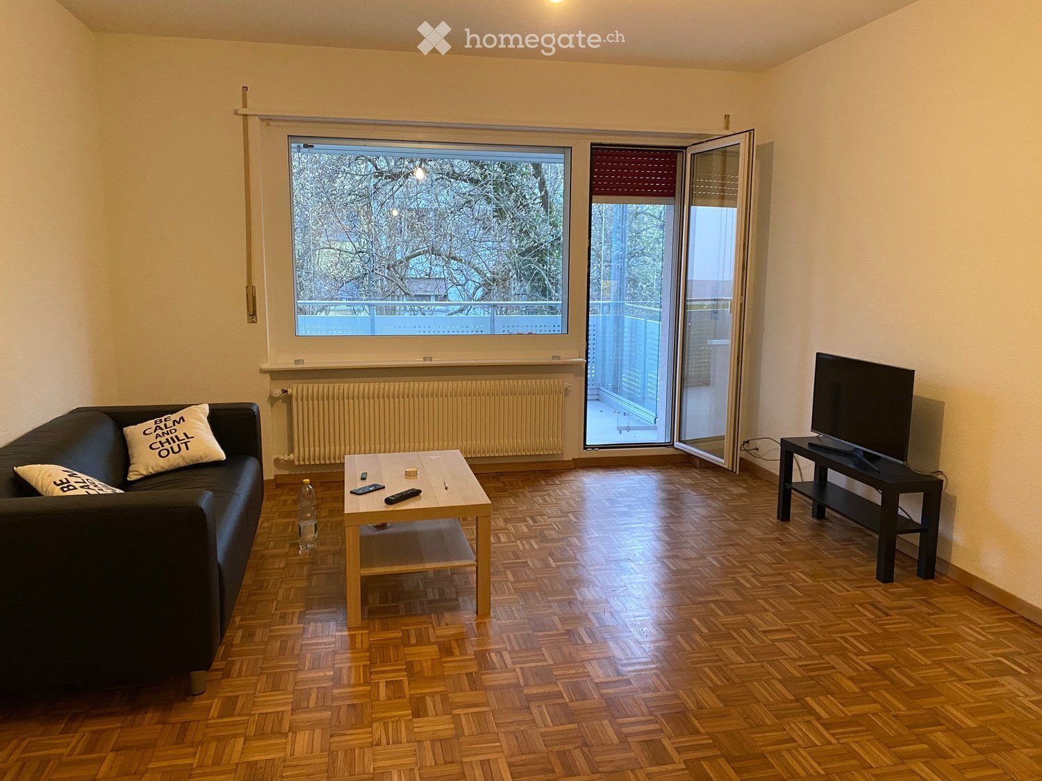 Living room with wooden parquet flooring, large window, sliding glass door to balcony, sofa with decorative pillow, coffee table, TV stand, and remote controls.