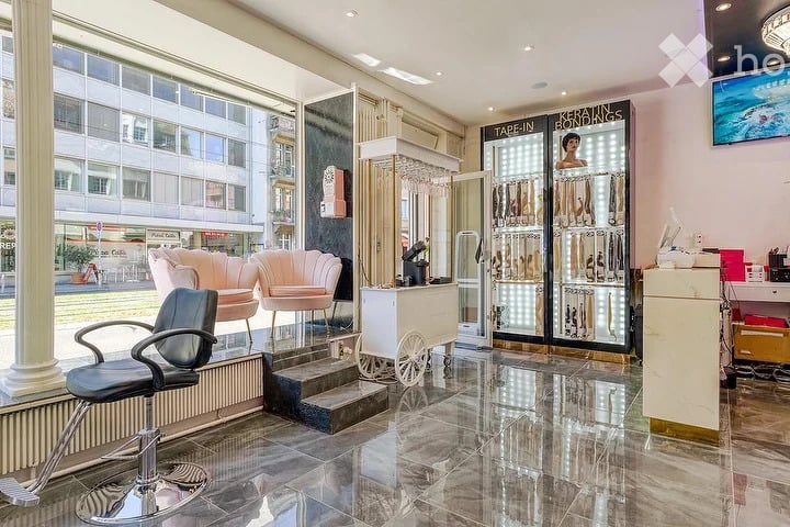 A modern, well-lit salon with a polished marble floor, a sleek black chair, a pink vanity stool, a large mirror, and a display cabinet with hair extensions.