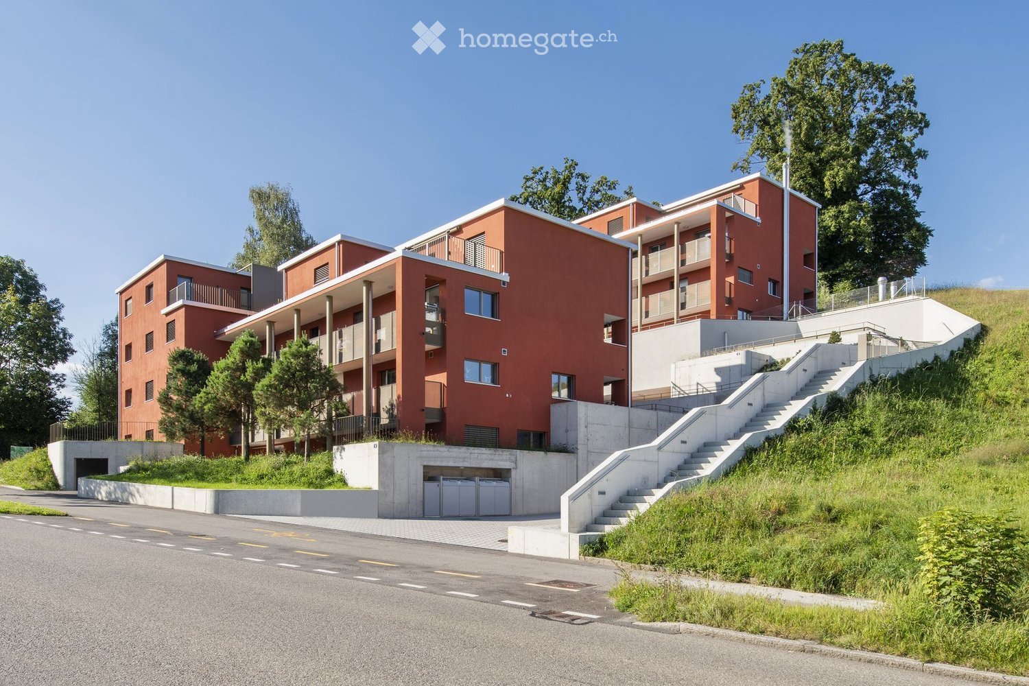 Modern multi-story residential building with red brick facade, balconies, and a raised ground floor entrance connected by concrete stairs.