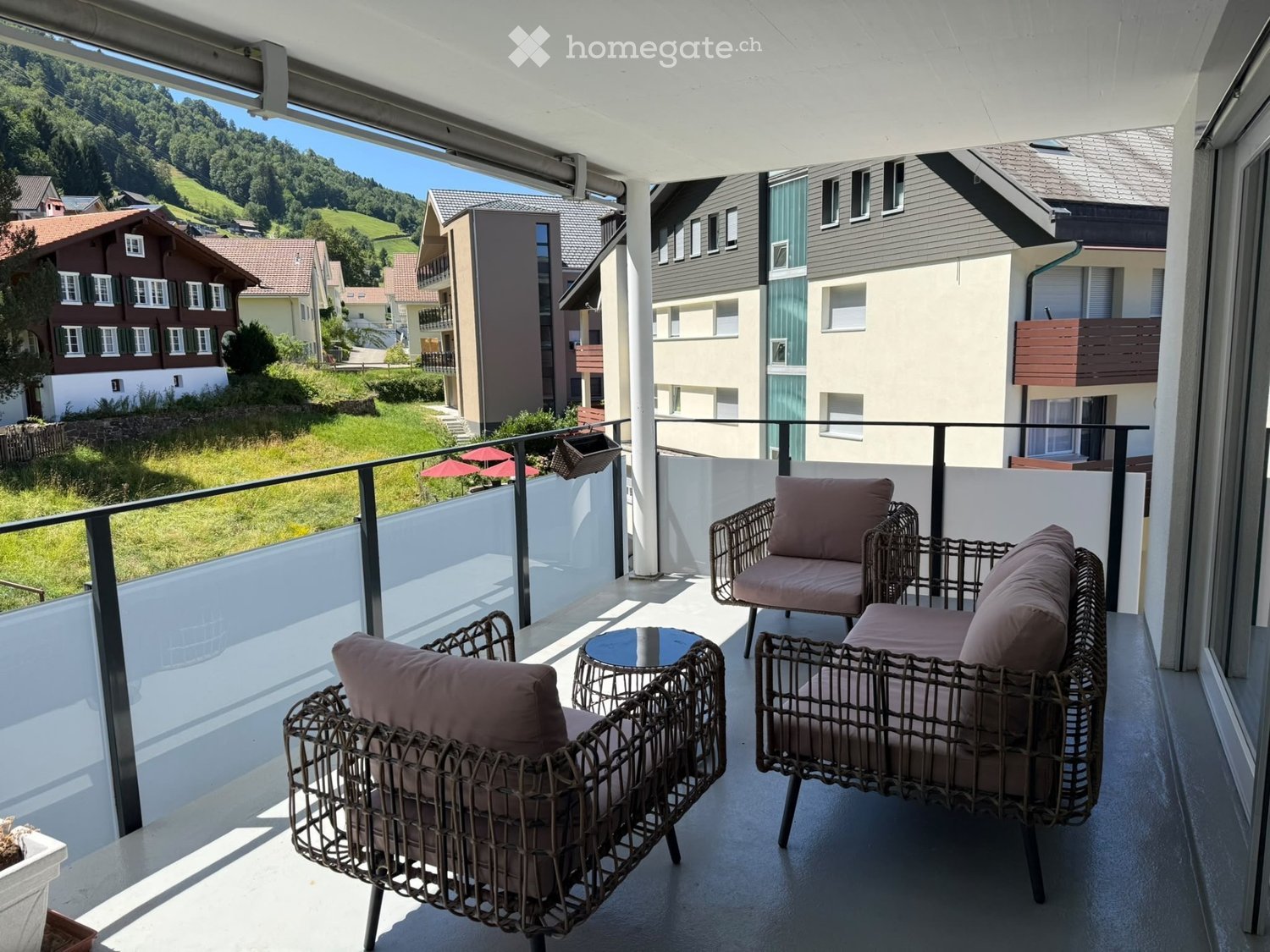Balcony with wicker chairs, sofa, and small table, overlooking a scenic town and green landscape.