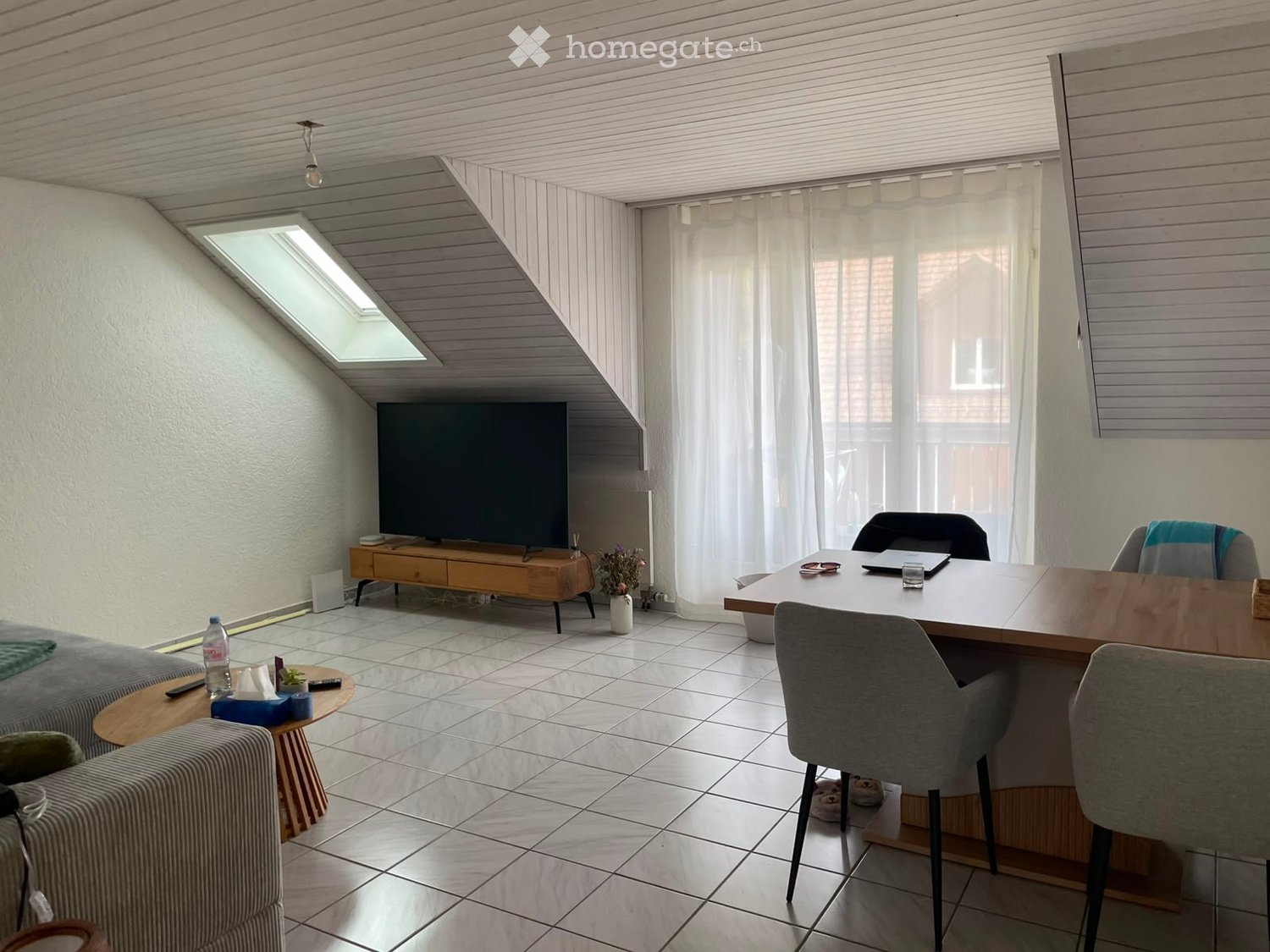 Modern living room with a sofa, coffee table, TV, and a dining area with a table and chairs. The room features large windows with white curtains and a skylight, creating a bright and airy atmosphere.