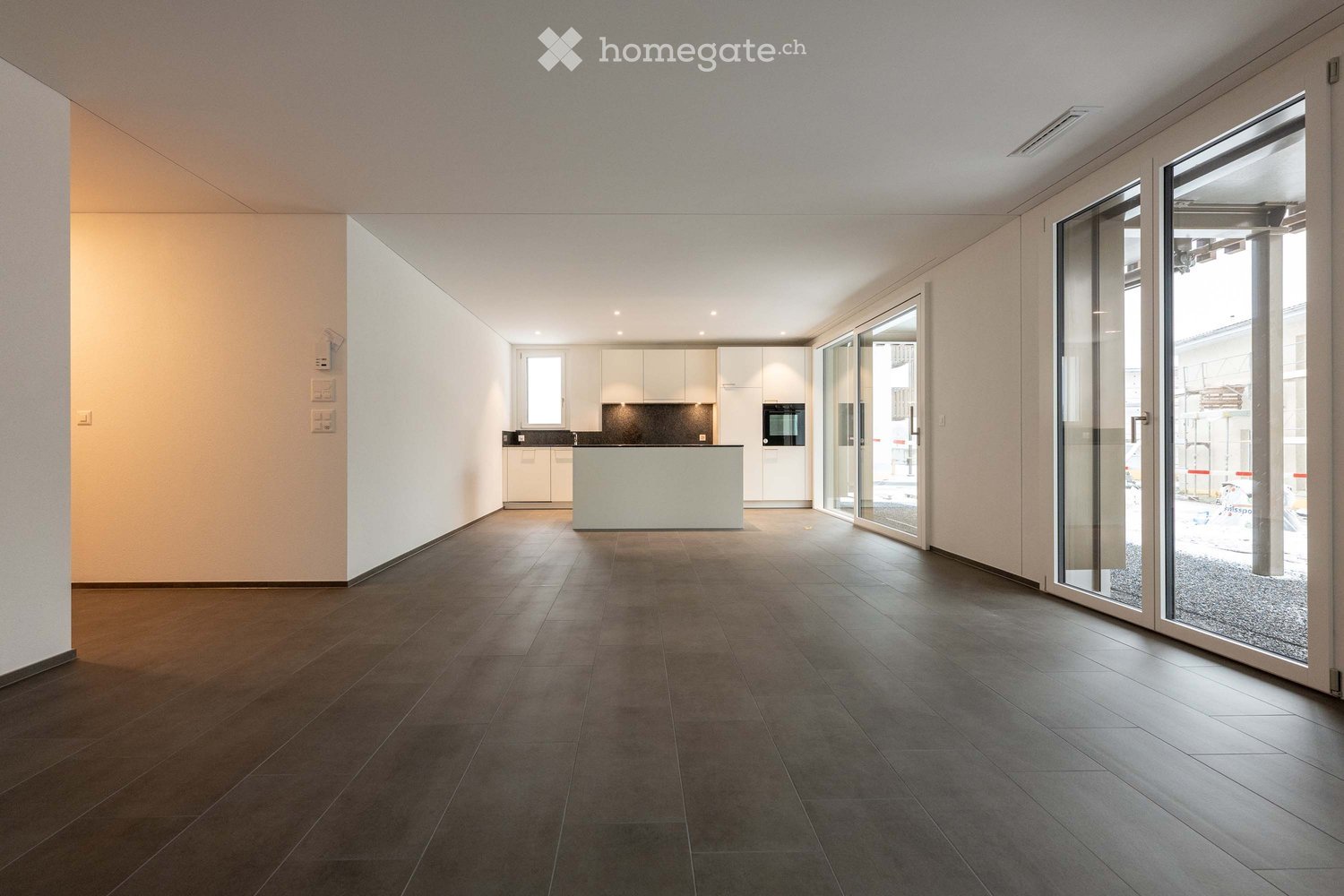 Open-plan unfurnished room with dark tiled flooring, white walls, and large glass sliding doors leading to an outdoor area. The room features a small kitchen area with white cabinets and a countertop.
