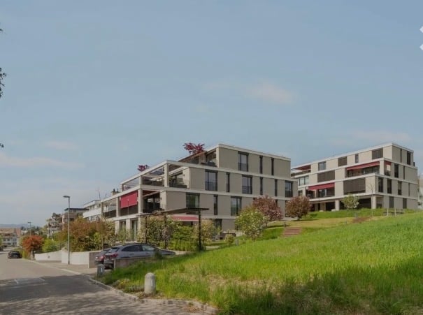 Modern multi-story residential building with balconies, surrounded by greenery and a paved pathway.