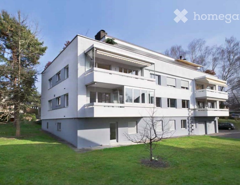 Modern white multi-story building with multiple balconies and large windows, surrounded by a well-maintained lawn and trees.