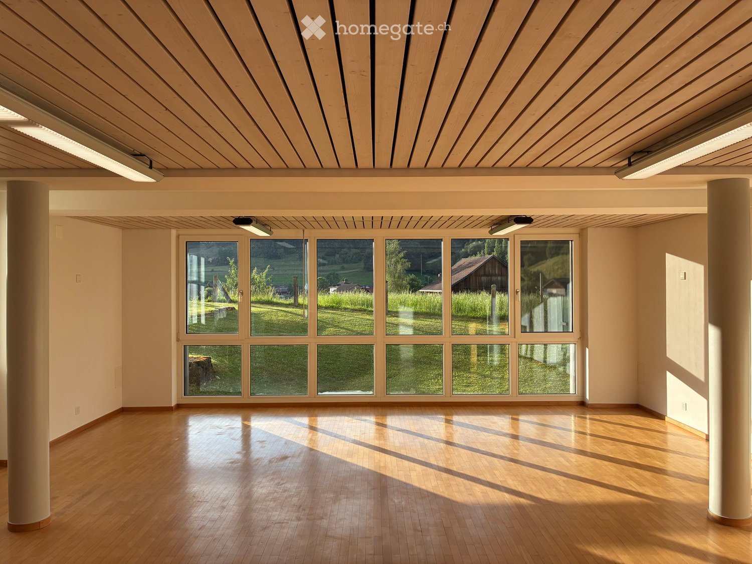 Empty room with wooden flooring, large windows, and a slatted wooden ceiling. Natural light streams in, illuminating the space.