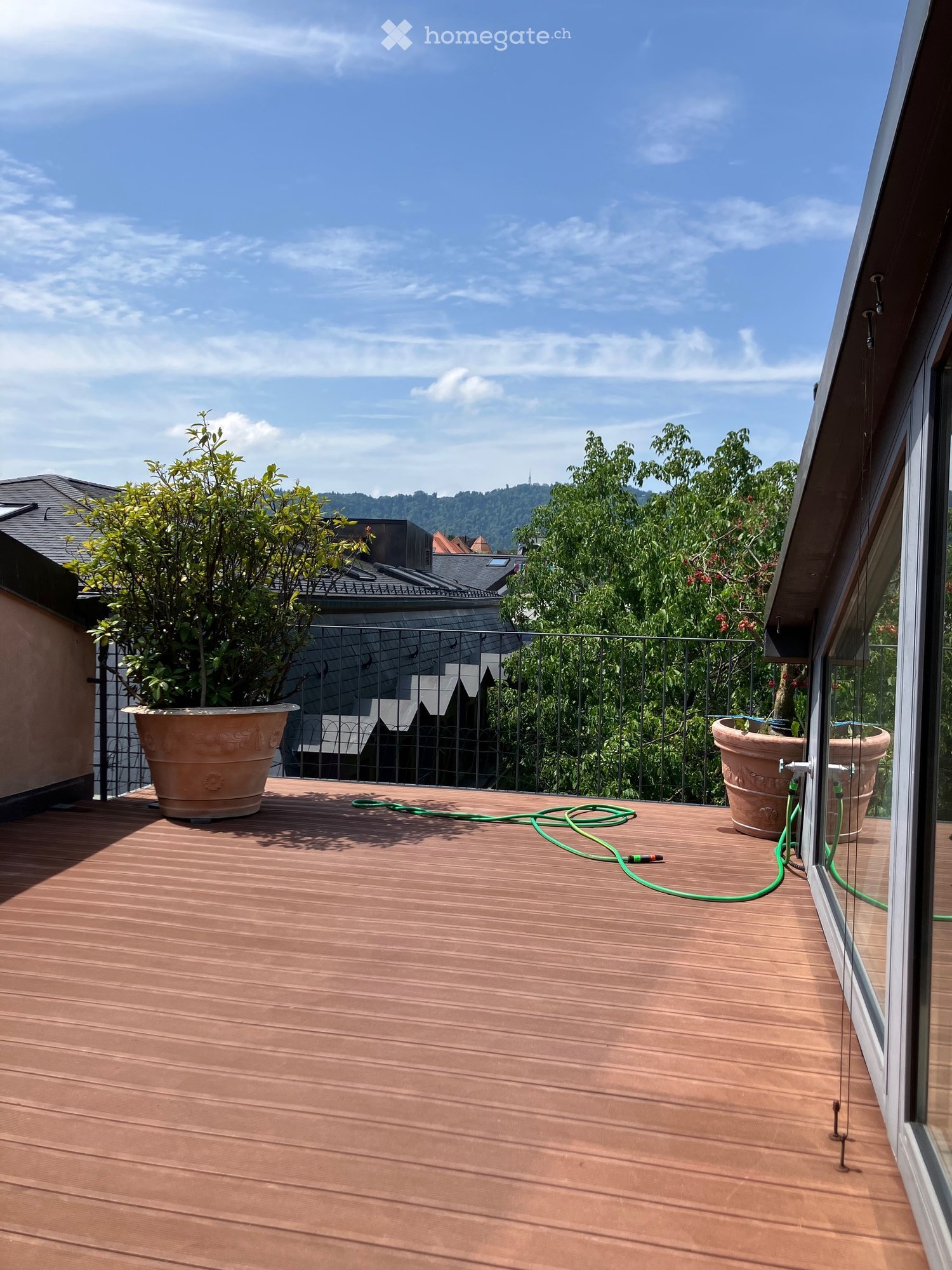 Wooden deck with two large pots, green hoses, and a metal railing overlooking a cityscape with trees and rooftops.