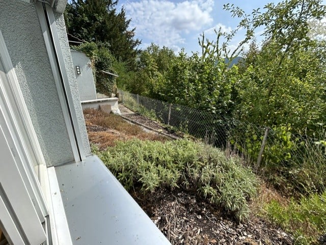 Balcony with a view of a fenced garden area, featuring some low plants and shrubs, with a concrete railing and a small staircase leading down.