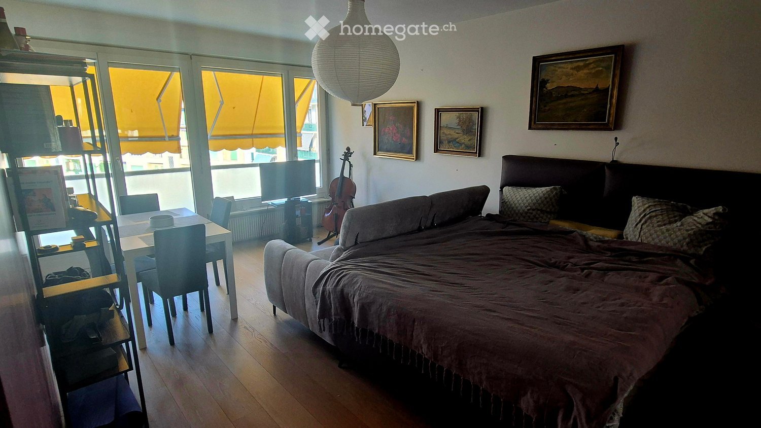 A cozy, well-lit room with a bed, sofa, dining table, chairs, a cello, and a television. The room features large windows with yellow blinds and several framed pictures on the wall.