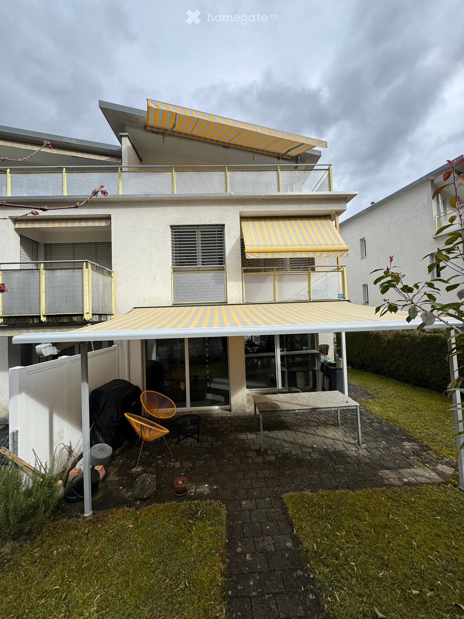 Modern two-story house with balconies, striped awnings, and a covered patio area with a table and chairs.