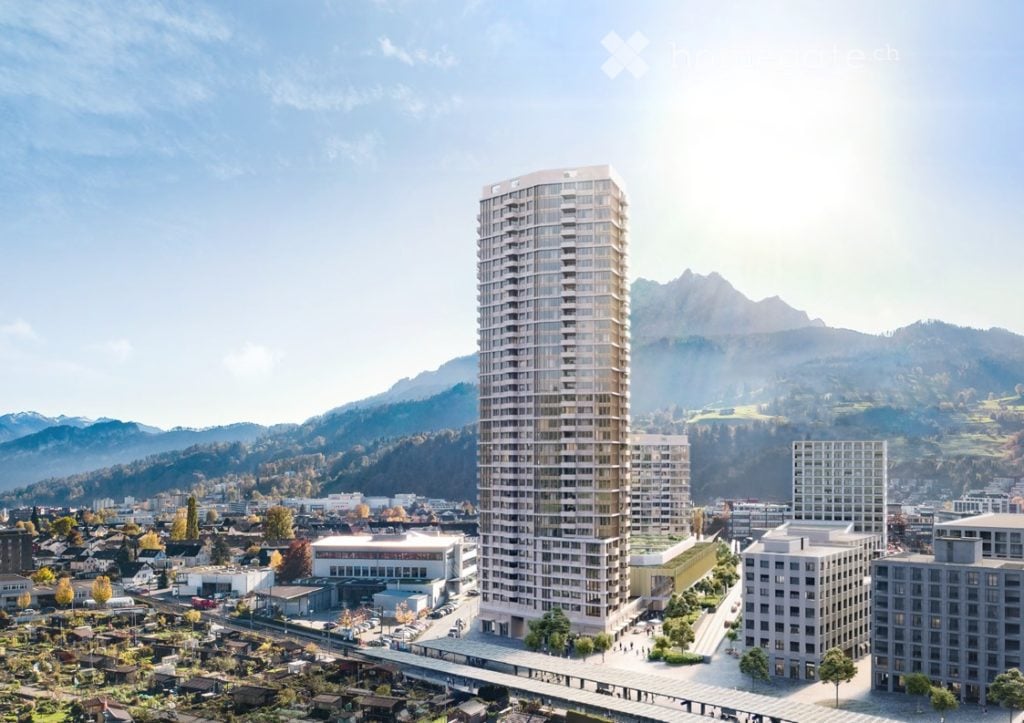 A modern high-rise building with reflective glass windows stands prominently in a cityscape, surrounded by smaller structures and a scenic mountainous backdrop.