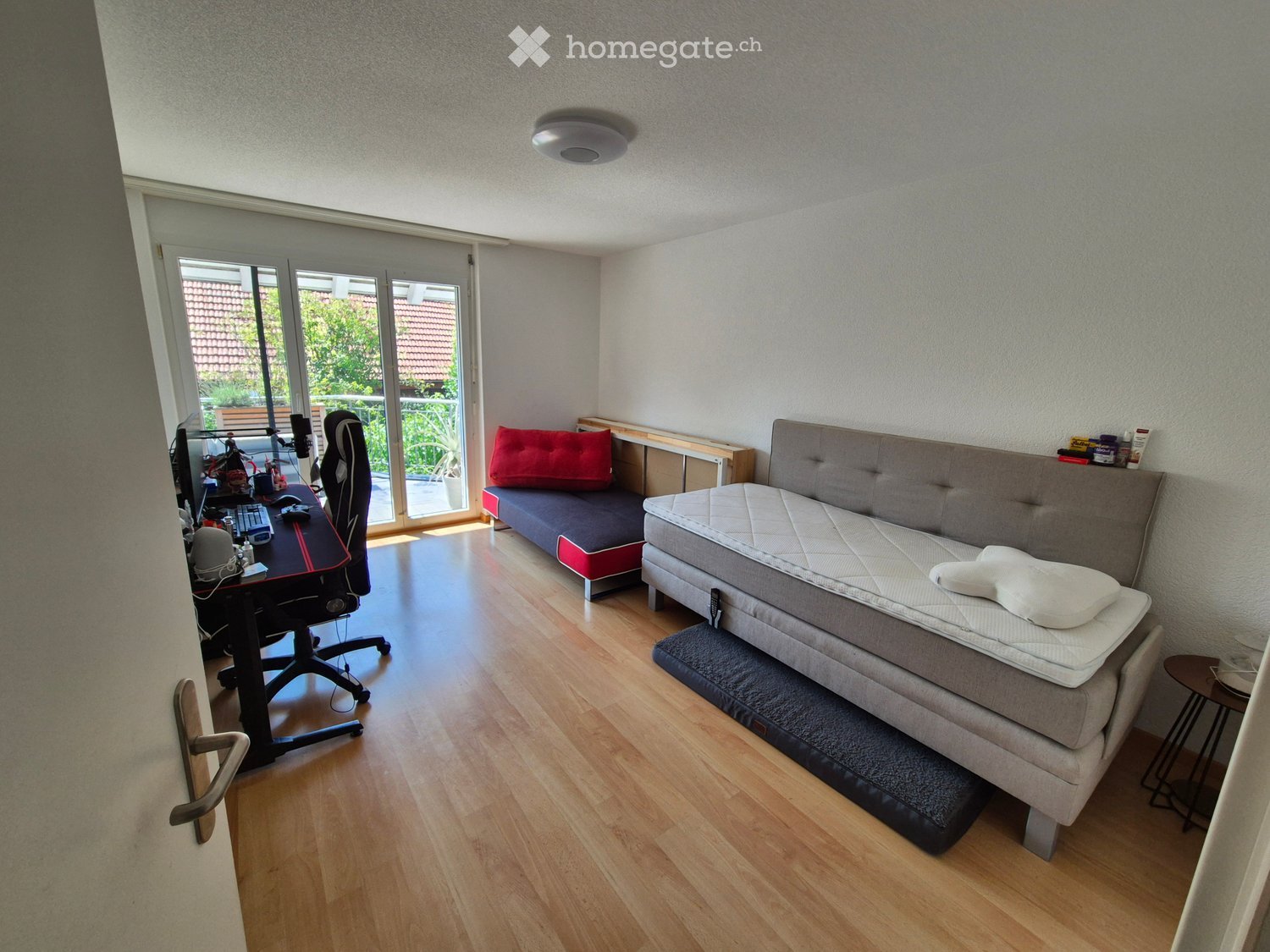A small, well-lit room with a wooden floor, a desk with a computer setup, a sofa with a red pillow, and a mattress on a platform bed.