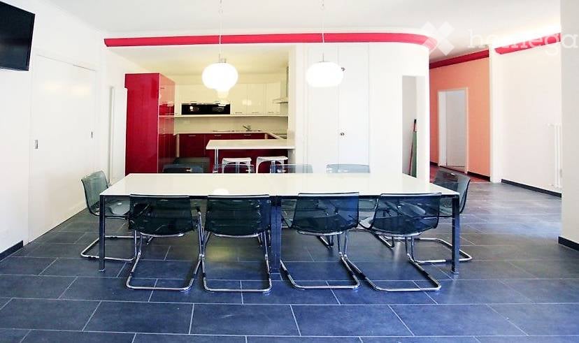 Bright, modern room with a large white table surrounded by six black chairs. In the background, a red kitchen area with cabinets, a sink, and a faucet is visible. The room features white walls, a red accent wall, and pendant lights.