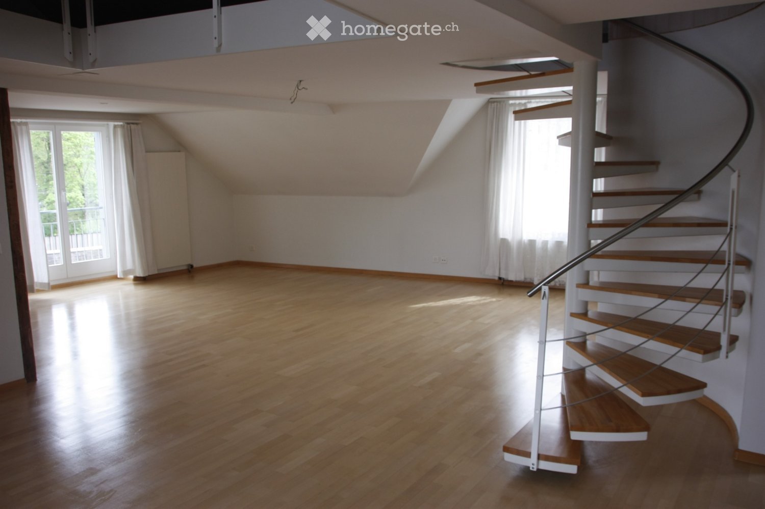 Empty room with wooden flooring, white walls, and a curved staircase leading upwards. Large windows with white curtains allow natural light to fill the space.