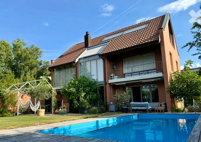 Two-story house with red brick exterior, large windows, balcony, and swimming pool in the garden.