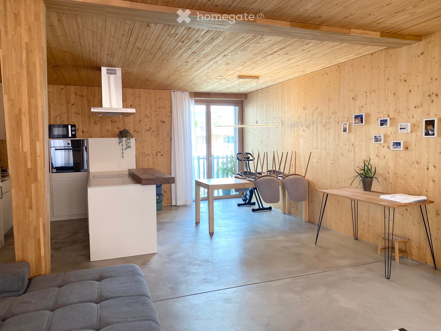 Open-plan living and kitchen area with wooden walls and ceiling, grey sofa, kitchen island, oven, refrigerator, dining table, chairs, and potted plants.