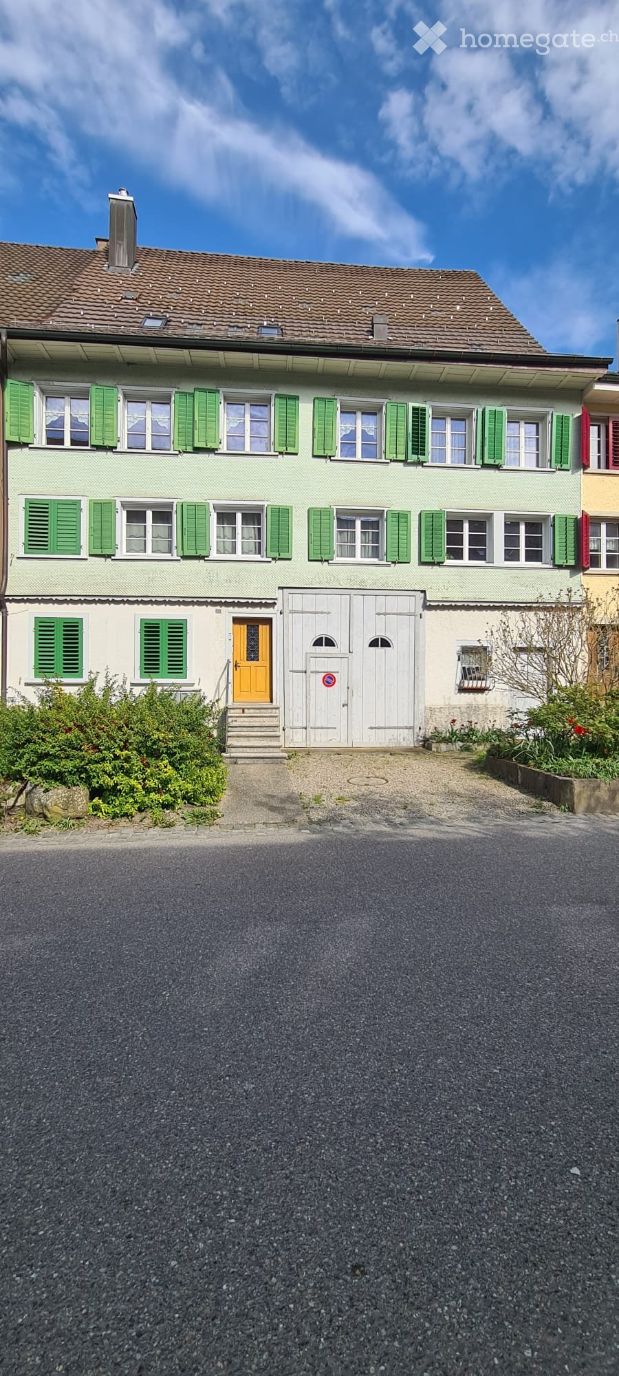 Two-story house with light green siding, green shutters, and a red-tiled roof. Features a white garage door, orange front door, and small garden area.
