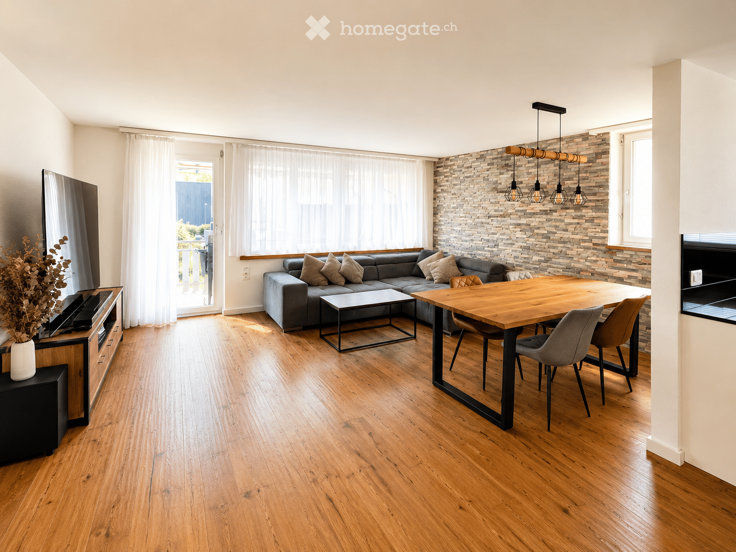 Modern living and dining area with wooden flooring, grey sectional sofa, wooden dining table with chairs, TV, and decorative vase with dried flowers.