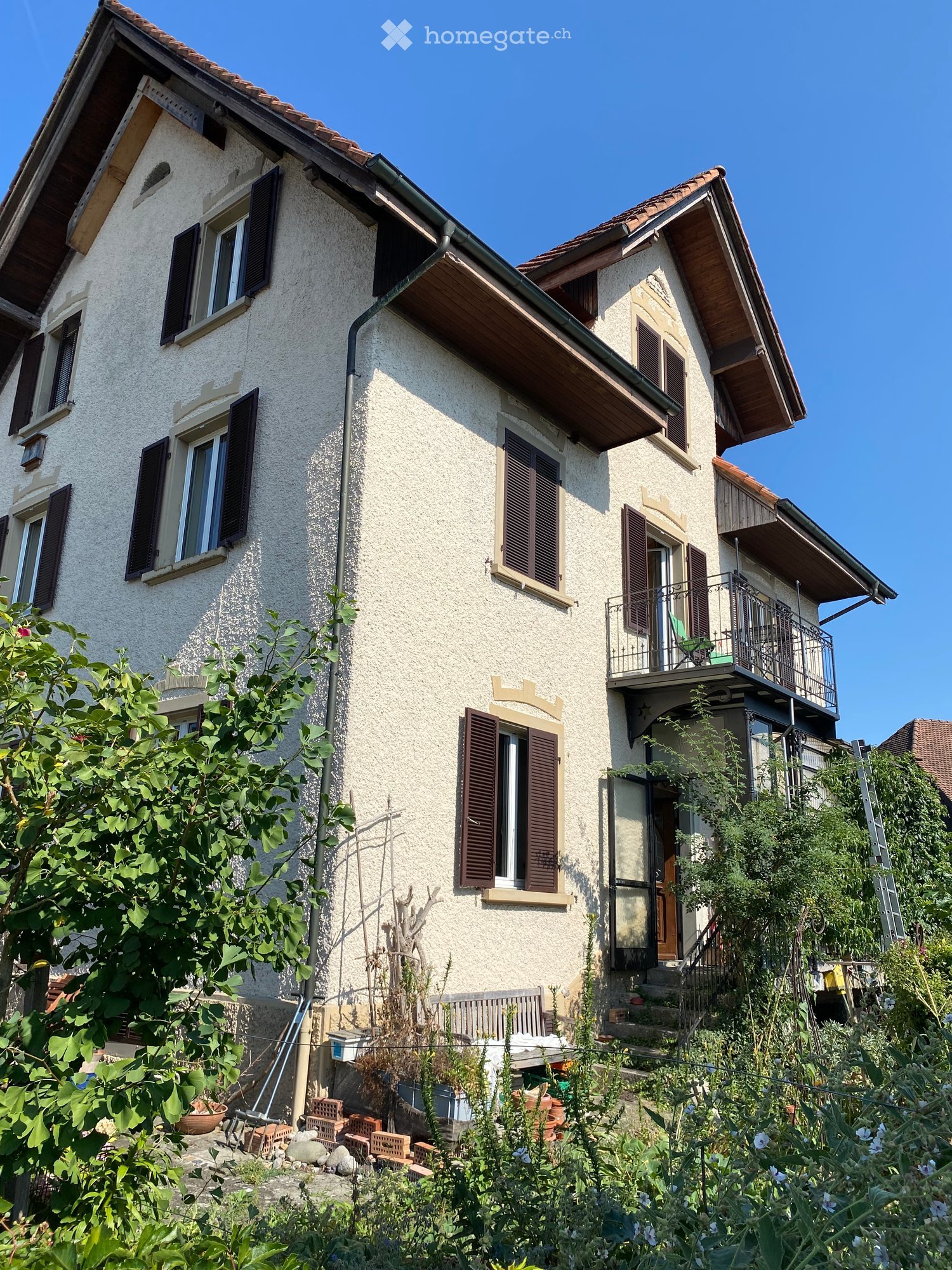 Two-story house with beige stucco exterior, multiple windows with dark shutters, a balcony on the upper floor, and a small garden area with plants and a bench.