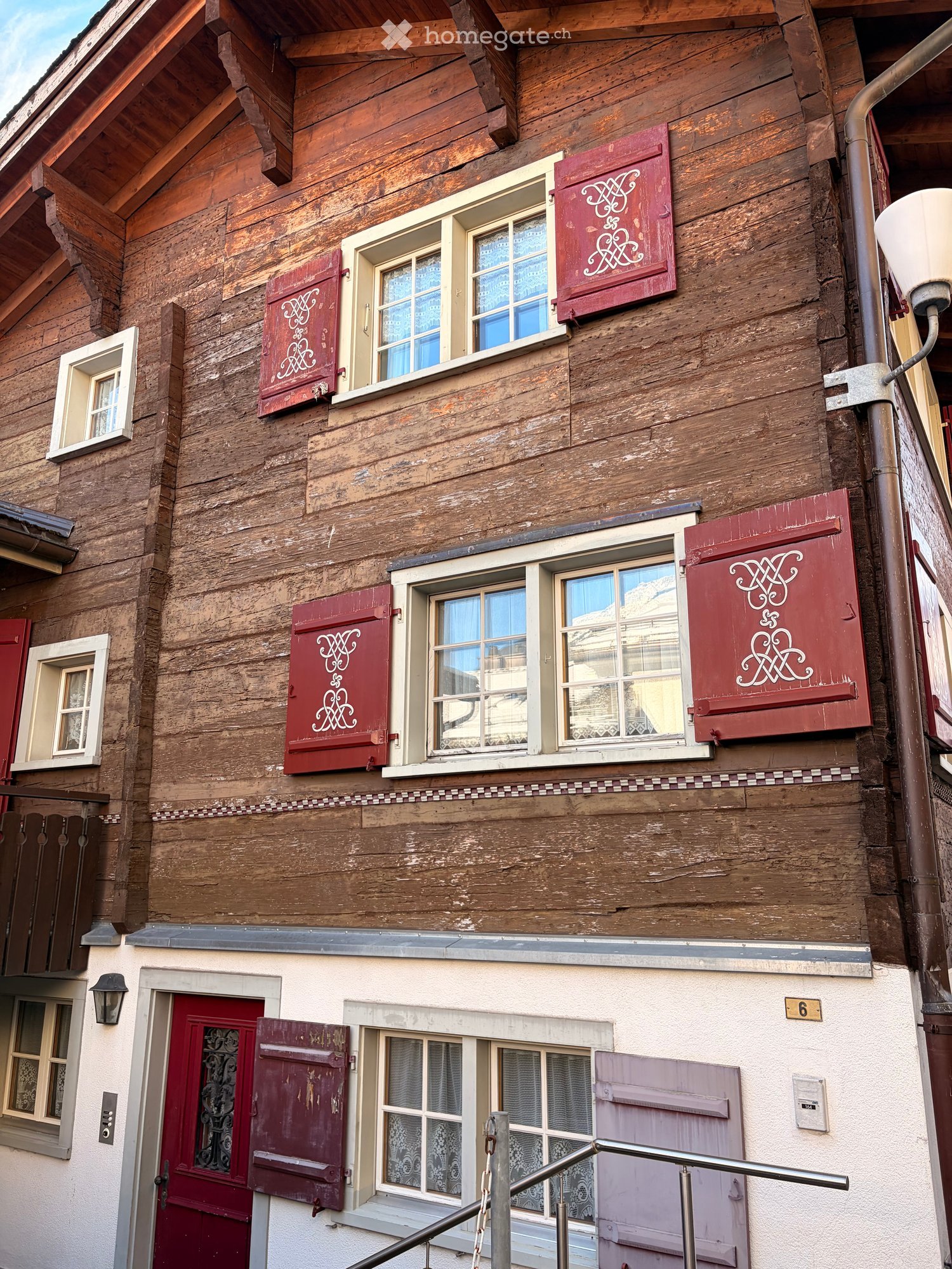 Two-story wooden building with red shutters, white-framed windows, and a decorative pattern on the shutters. The lower level has a white facade with a red door and a small balcony.