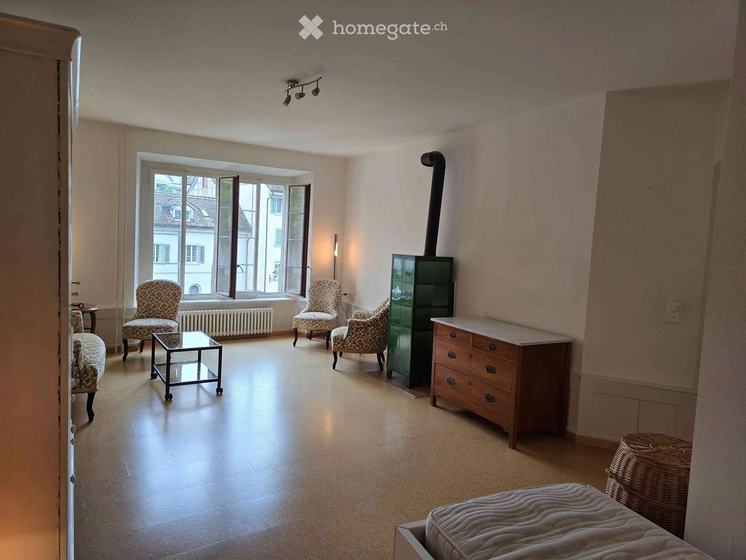 A well-lit room with a mix of modern and classic furniture, including a sofa, coffee table, chairs, a small table, and a drawer unit. The room features large windows with a view of the outside and a radiator beneath them.