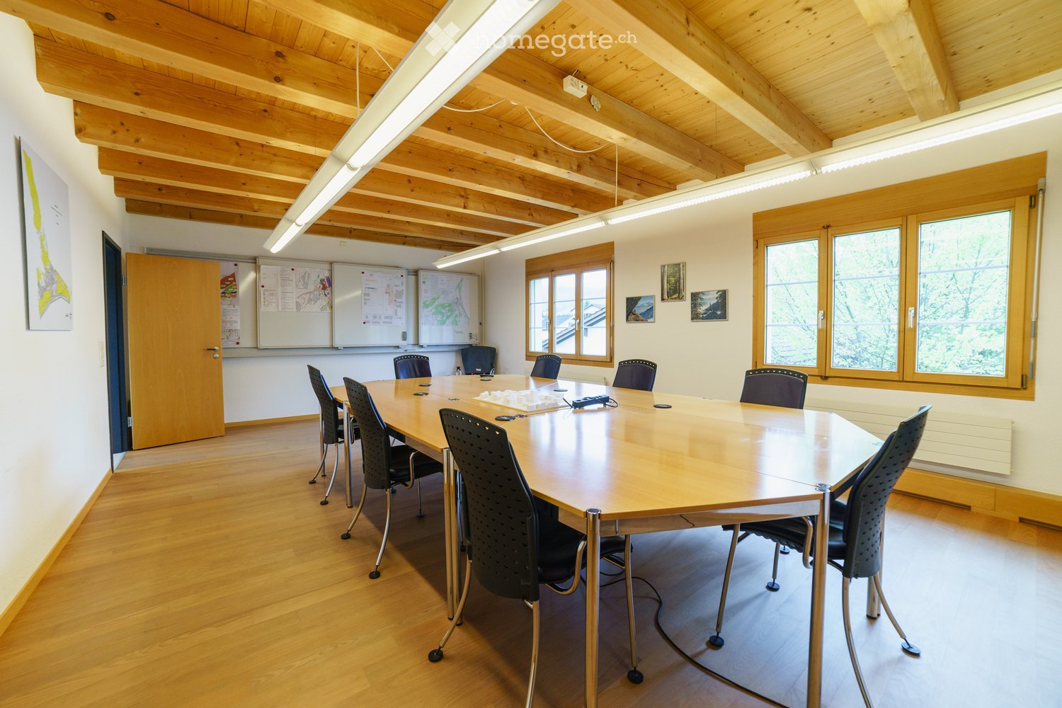 Bright conference room with wooden ceiling beams, large windows, a long wooden table surrounded by black chairs, and a whiteboard area with maps and charts.