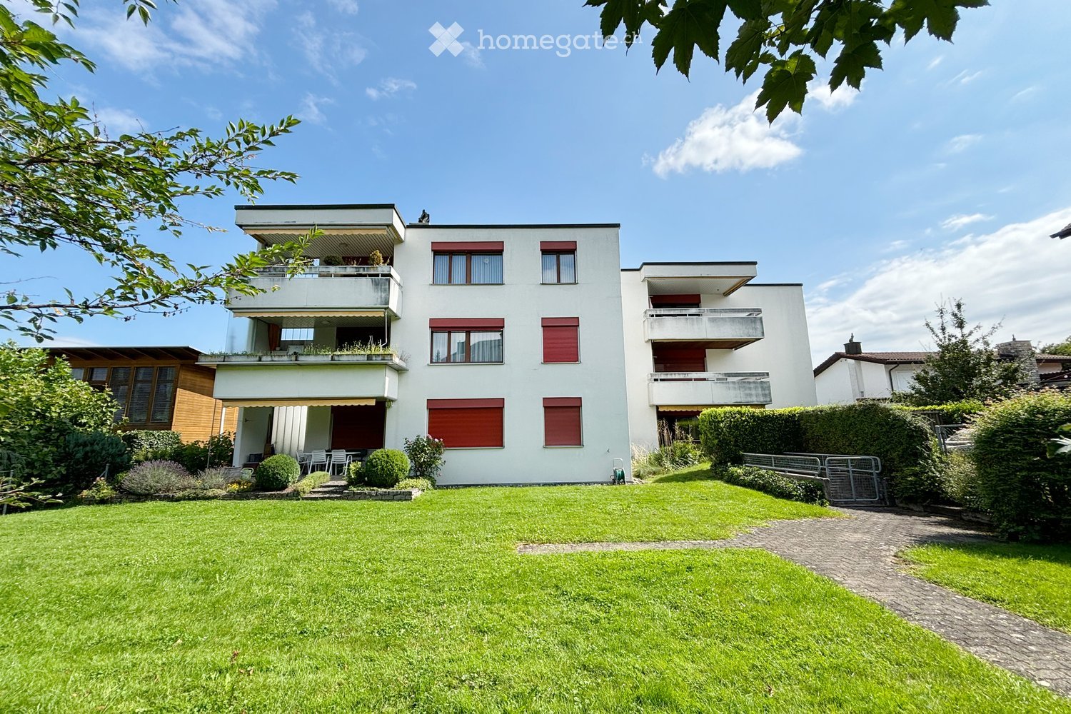 Modern two-story house with white exterior, red shutters, multiple balconies, and a well-maintained lawn in front.