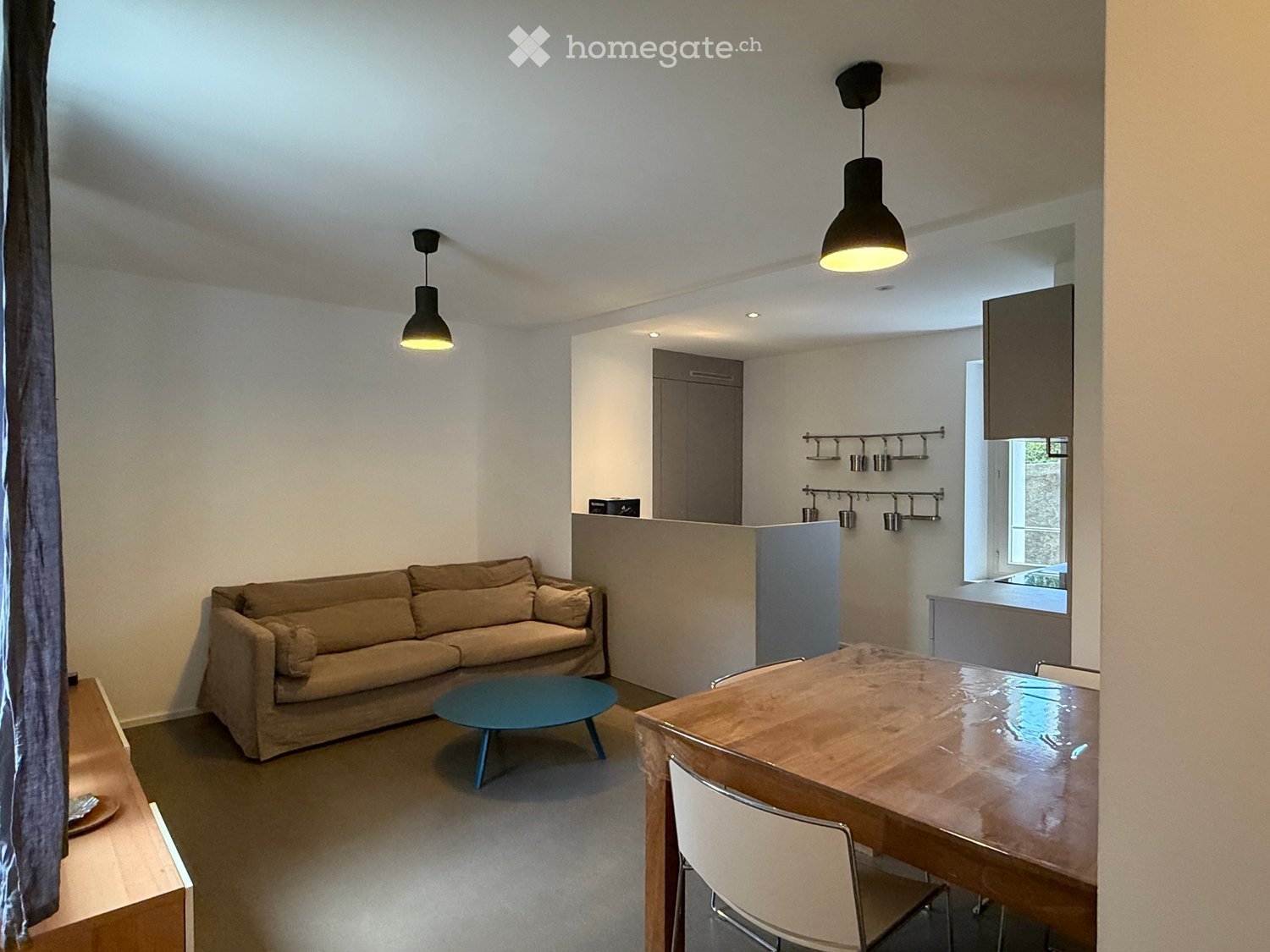 Open-plan living and dining area with a sofa, coffee table, dining table with chairs, and a small kitchen area with cabinets and pendant lights.
