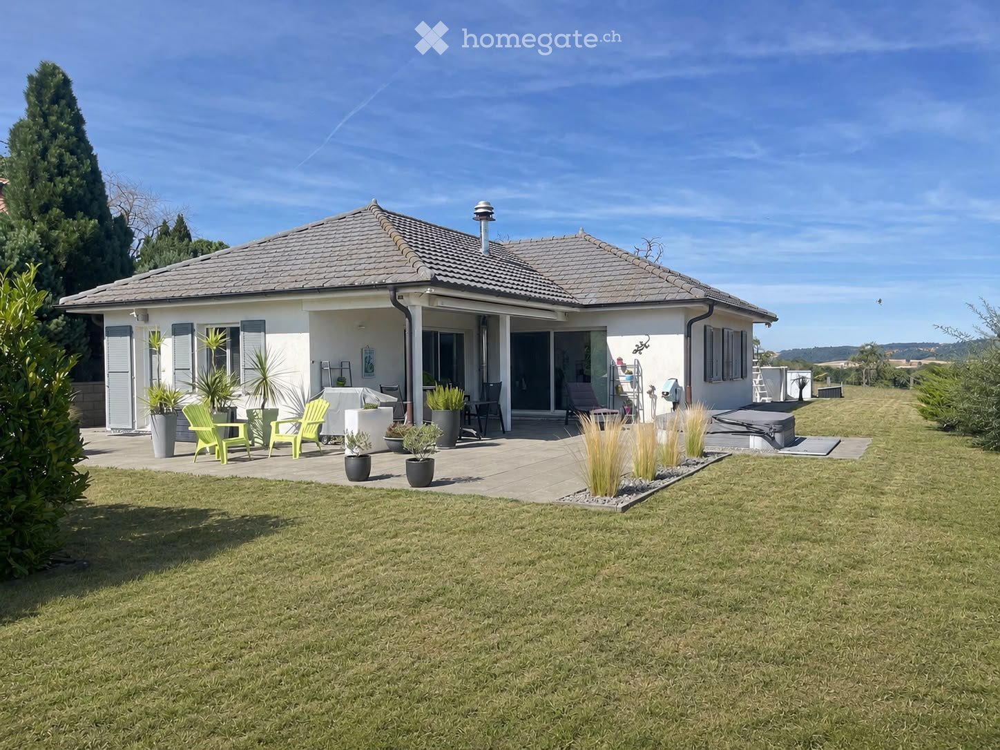 A modern single-story house with a tiled roof, surrounded by a well-maintained lawn and garden. The patio area features green chairs, tables, potted plants, and a small hot tub.