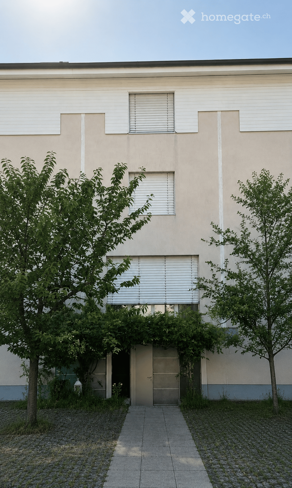 Modern single-story building with light beige facade, multiple windows with white blinds, and a central entrance door surrounded by greenery and trees.
