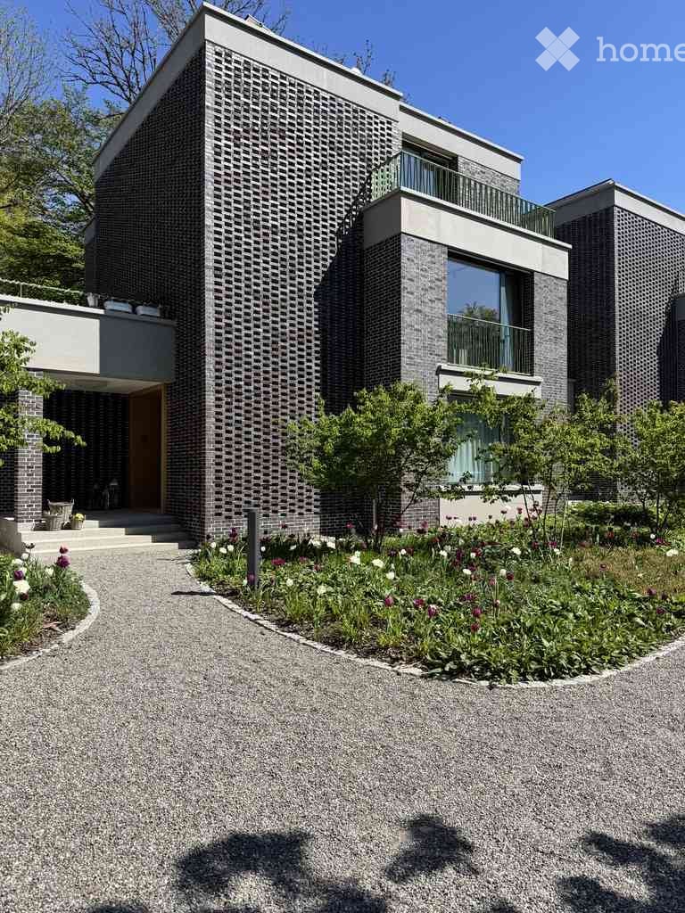 Modern two-story house with grey brick facade, large windows, and balconies. The front features a landscaped garden with flowers and a gravel driveway.