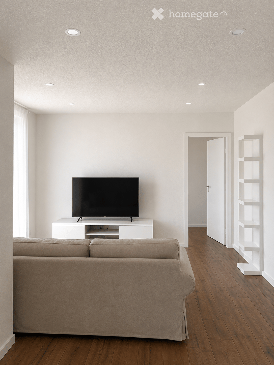Modern living room with a beige sofa, flat-screen TV on a white stand, wooden flooring, and white shelving unit.
